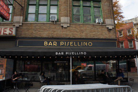 The exterior view of Bar Pisellino. The storefront features outdoor seating space with a black awning decorated with the text “BAR PISELLINO” in white. Above the awning is a black plaque against which gold text that reads “BAR PISELLINO” is printed.