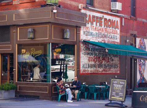 The diagonal view of Caffé Roma. There is an outdoor dining space with teal chairs, an awning and white tables. On the side is a mural with a white background and red text that reads “CAFFÉ ROMA Fine ITALIAN PASTRIES.”
