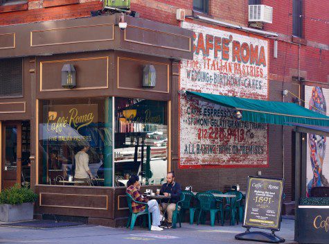 A diagonal view of Caffé Roma.  There is an outdoor dining area with blue chairs, an awning and white tables.  On the side is a mural with a white background and red text that reads 