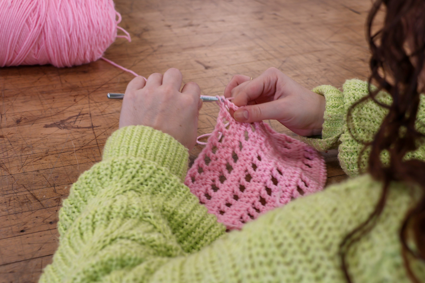 Becca Panes wears a neon green knitted sweater. Becca holds a metallic crochet hook and pink yarns.