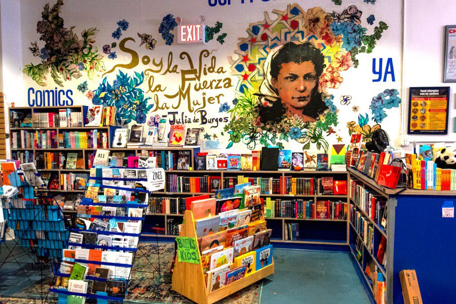 An interior view of the Bluestocking bookstore. In the foreground are shelves of comic books. On the wall is a painting of Julia de Burgos in brown. On the wall is text that reads “Soy La vida, La Fuerza, La Mujer,” which translates in English to “I am Life, Force and Woman.”