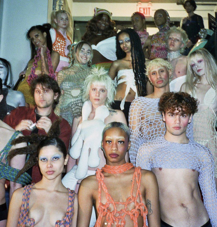 A large group of people wearing knit-themed and high-fashion outfits stands on a staircase.