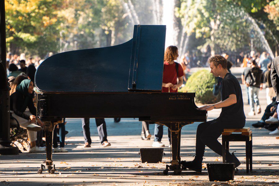 Colin Huggins wearing all black playing a grand piano in the middle of a park. The piano’s paint is worn off on the edges and text “this machine kills fascists” is etched onto the paint in all caps.