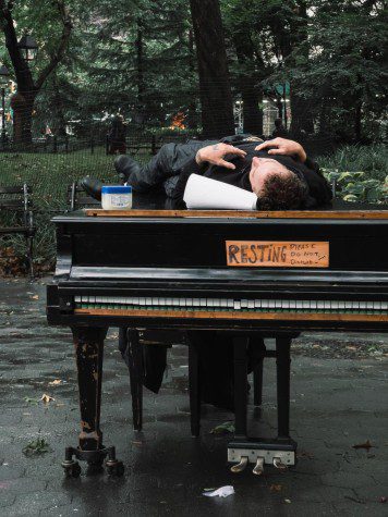 Colin Huggins wearing all black lying on top of a grand piano in Washington Square Park with his hands put on his chest. Next to his head is a paper towel roll. A plaque is put on the piano cover with handwritten text “resting please do not disturb.”