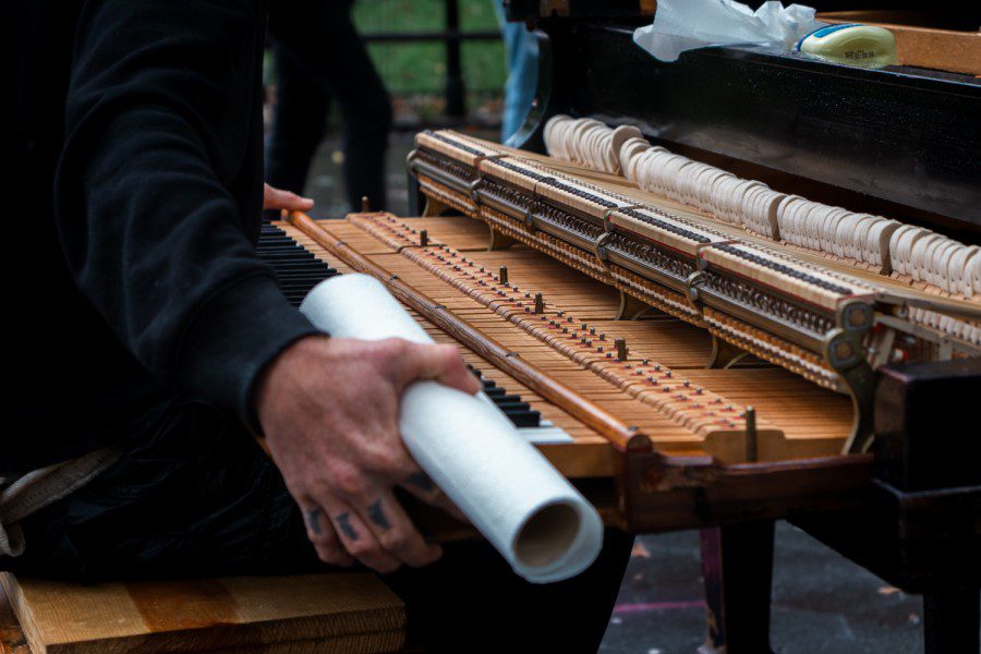 A pair of hands with tattoos on the fingers holding the keyboard of a grand piano. The piano is taken apart and exposing its internals. The right hand is also holding a paper towel roll.