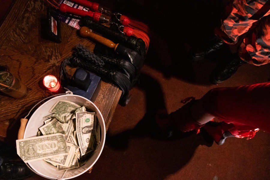 A close-up view of a wooden tabletop on which are various whips, a stamp, a red candle and a bucket of one-dollar bills.