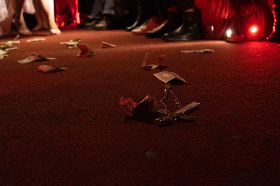 A silver cross and numerous crumpled one-dollar bills lay on the ground. The floor is lit in red.