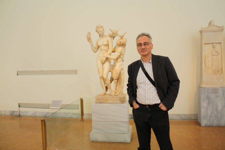 Professor Yunus Tuncel wearing a black suit jacket, a white shirt and black pants standing in front of a statue at an exhibition. He is smiling and wearing glasses.