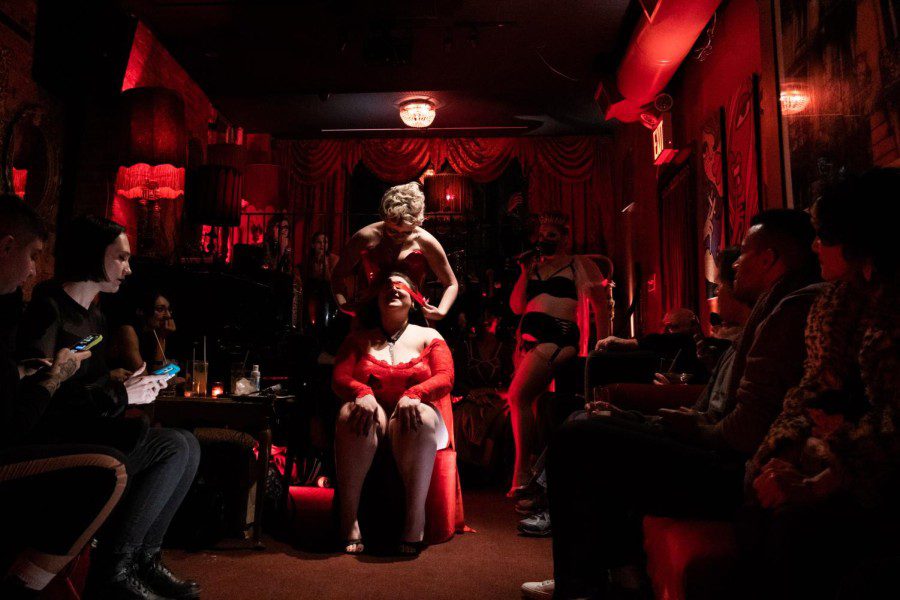 A performer in short golden hair wraps a red blindfold around the eyes of another female performer sitting down and wearing a red gown. The audience surrounds them on chairs in a room dimly lit by red lights.