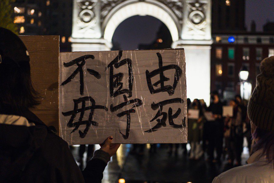 A person holding a sign that reads “不自由 毋宁死 (Give me freedom or give me death).” In the background is the Washington Square Arch lit up by lights.