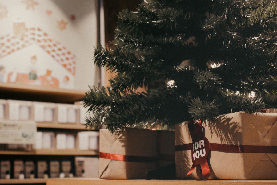 A small christmas tree placed on top of a wooden shelf with two gift boxes wrapped with brown paper under it. The boxes are tied with red straps with text “FOR YOU” in all caps. In the background are shelves of indistinguishable goods.