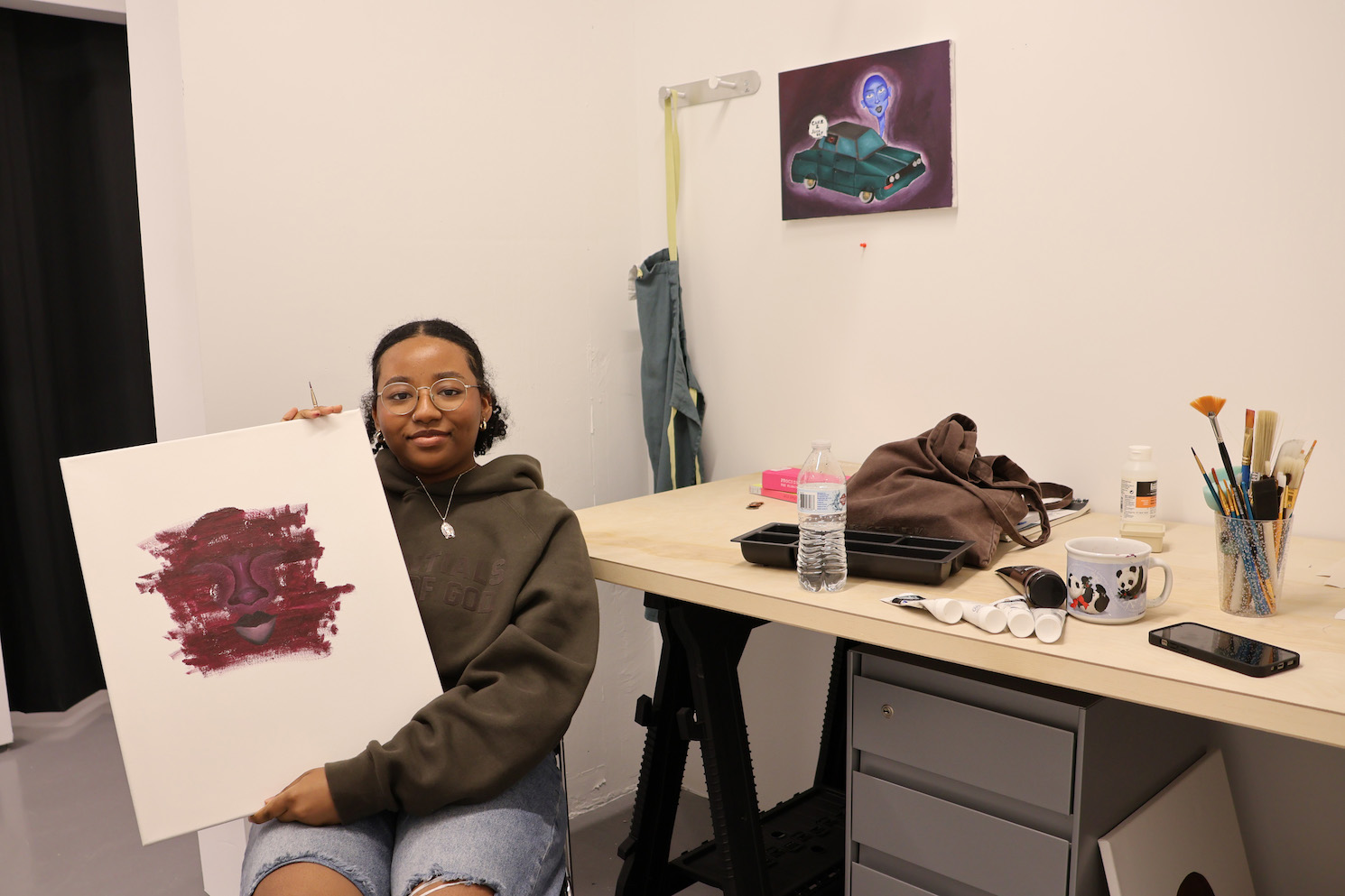 Keydi Alvarez, wearing a dark brown hoodie, holds a piece of painting. The painting depicts an illustration of a face in dark red. Behind Keydi is a desk with a dark gray drawer at the bottom. On the table are holder for paints and brushes and a brown tote bag.