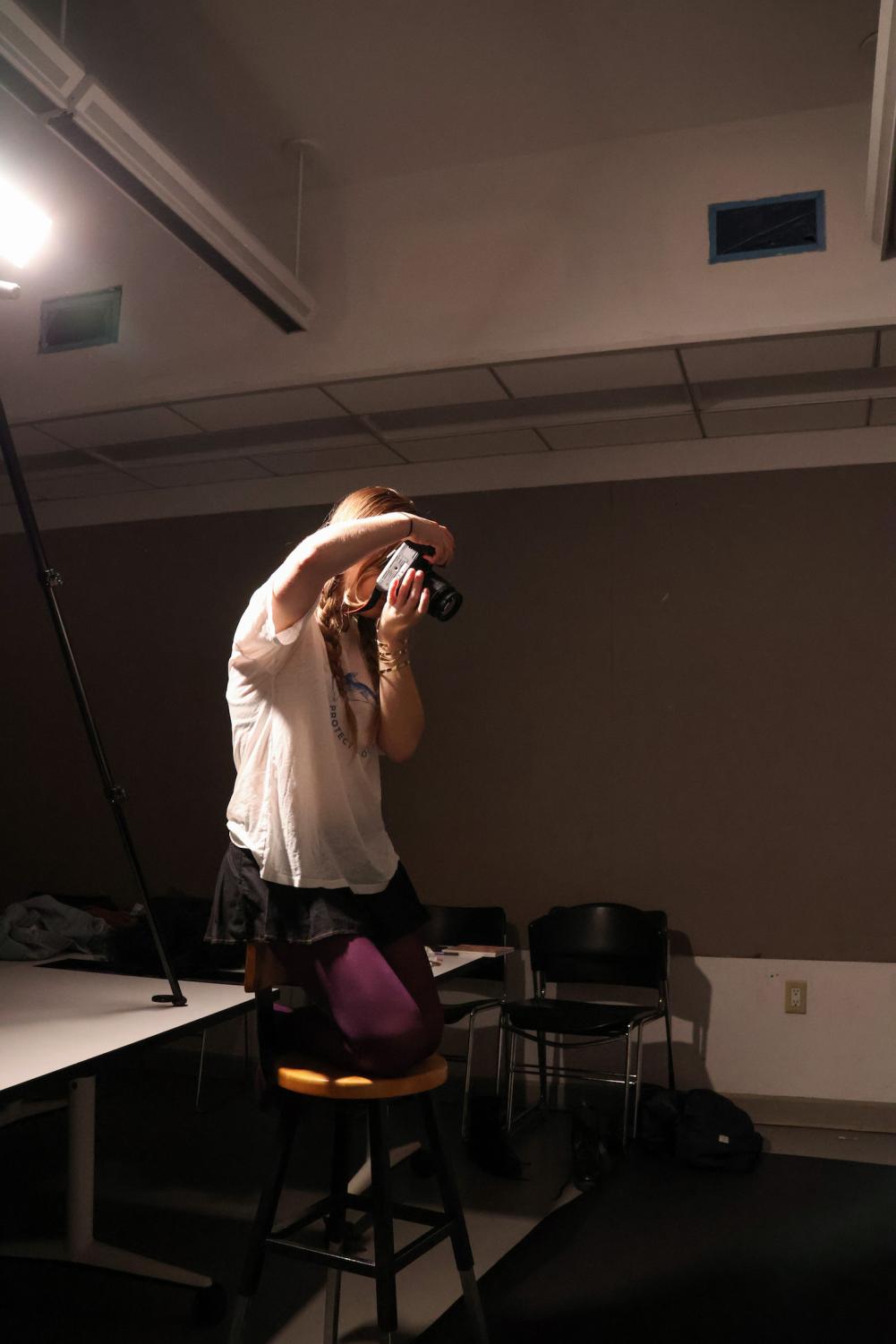 Photograph of a female college student with braided blond hair wearing a white t-shirt, a black skirt, maroon leggings, and gold bracelets. She kneels on a stool holding a DSLR camera up to her face pointing it.
