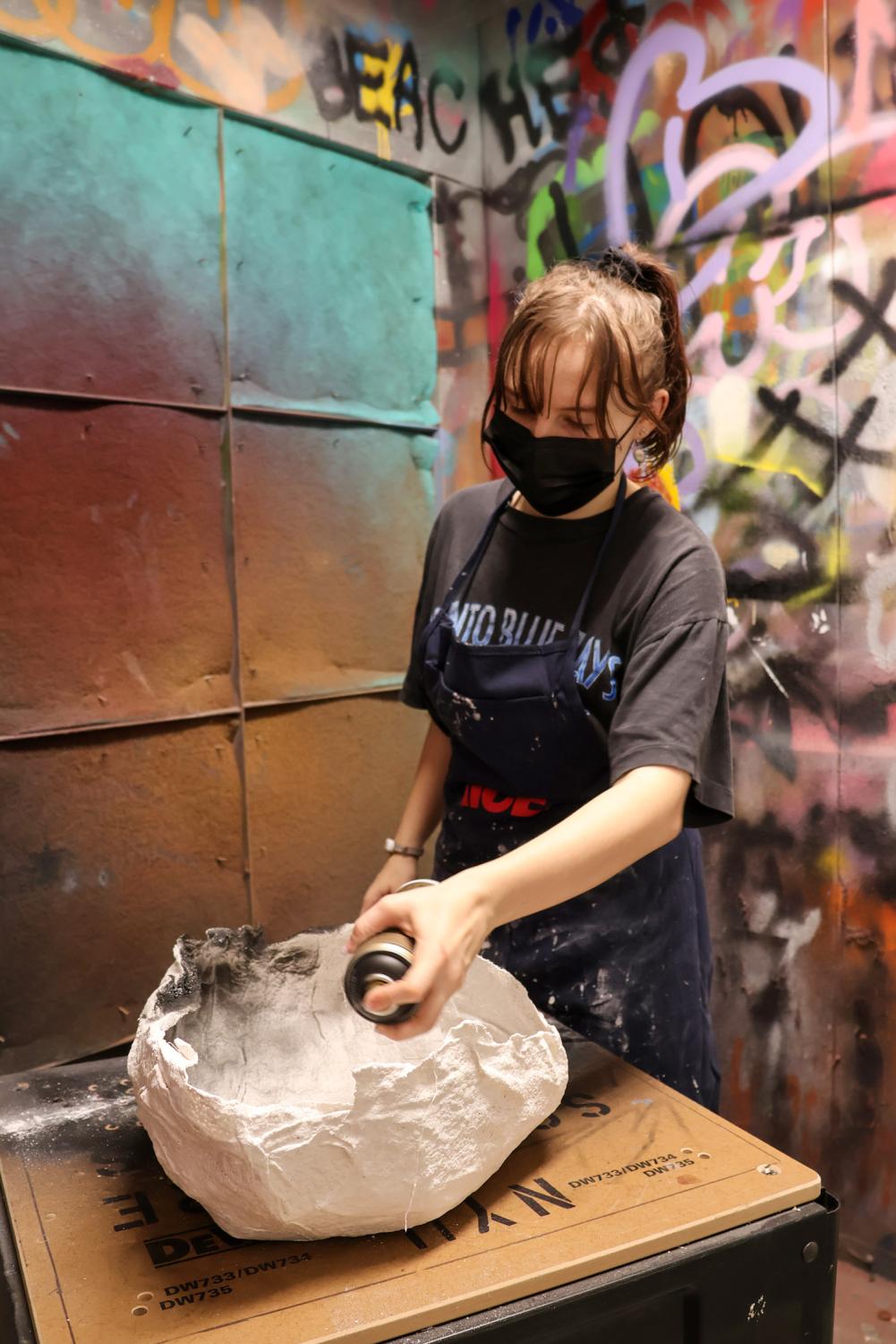 Sarah Gelleny wears a black mask, black t-shirt and a black apron. Sarah holds a bottle of black spray paint and sprays the paint onto a white mold on a wooden table.