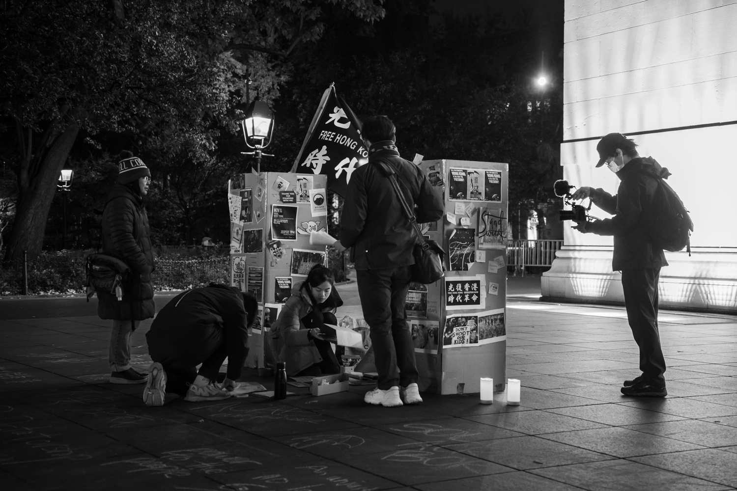 Four people gather around two columns made of cardboards that have several posters on them. One of the posters reads “FREE HONG KONG. REVOLUTION NOW” in all caps and traditional Chinese. There is another person holding a camera toward the group. Behind them is the Washington Square Arch. The photo is black and white.