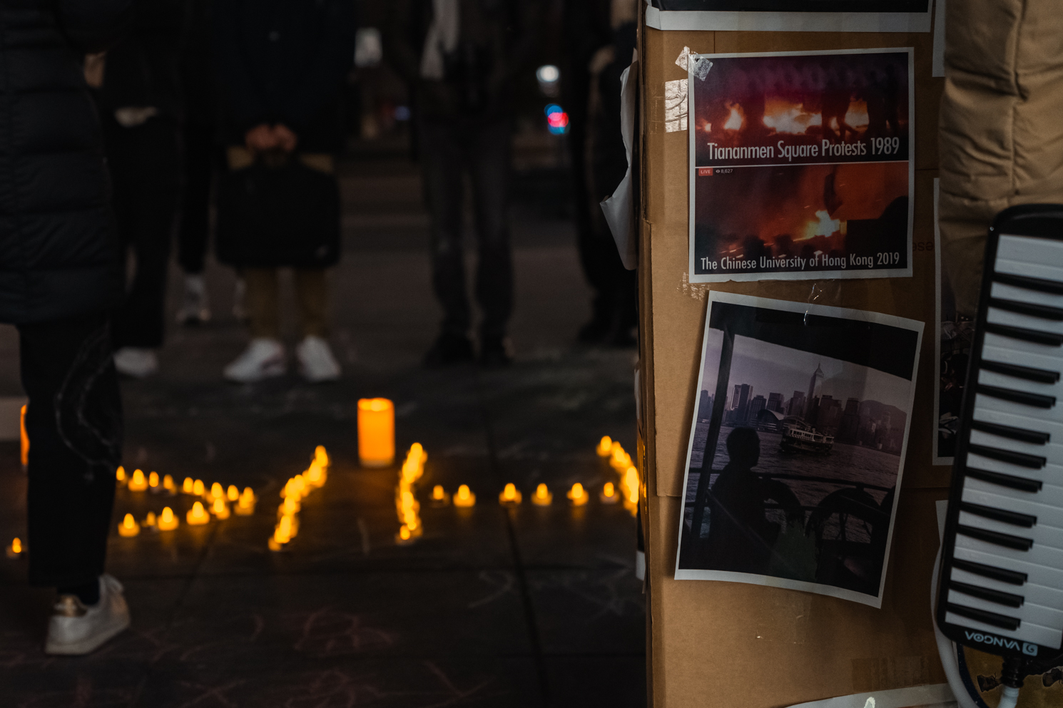 In the foreground are posters showing the Tiananmen Square Protest in 1989. In the background are candles on the ground in a formation that spells "HK"