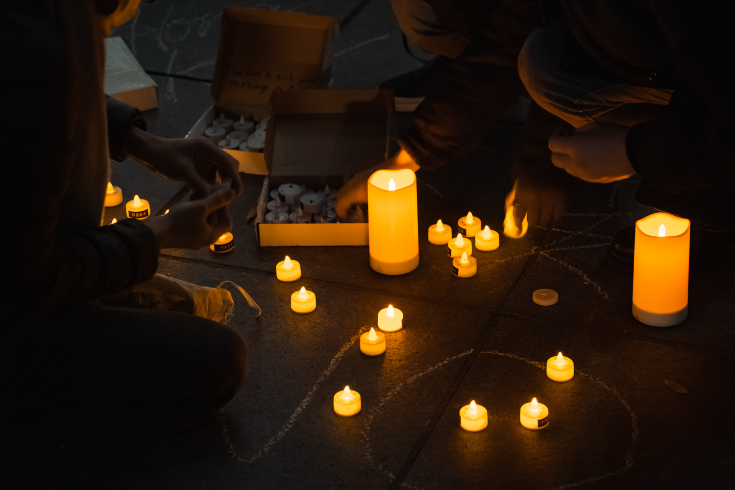 Three people squat close to the ground as they pick up small, candle-shaped lights from two boxes. In the middle are several candle-shaped lights turned-on, and two larger candle-shaped lights. There are chalk writings on the ground. Some of the candle-shaped lights have black labels on them.