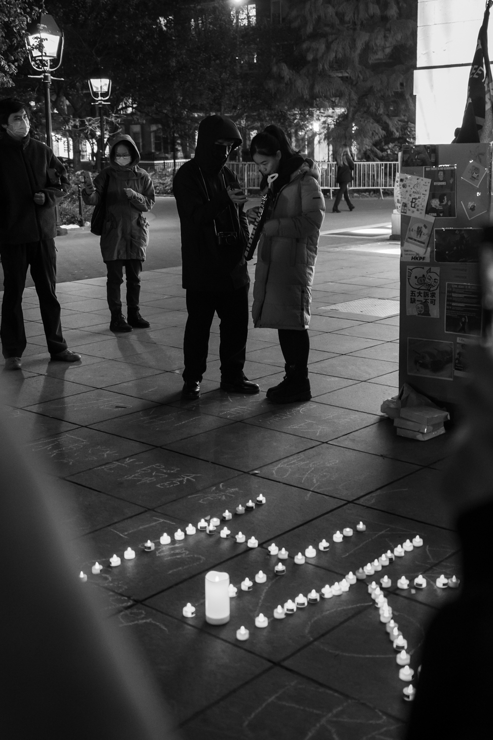 A female plays a melodica while looking at a phone held by another person wearing a hoodie and a black mask. Next to them is a column made of cardboard, with posters hung on it. In front of them are the letters “H.K.” laid out with candle-shaped lights. The photo is black and white.