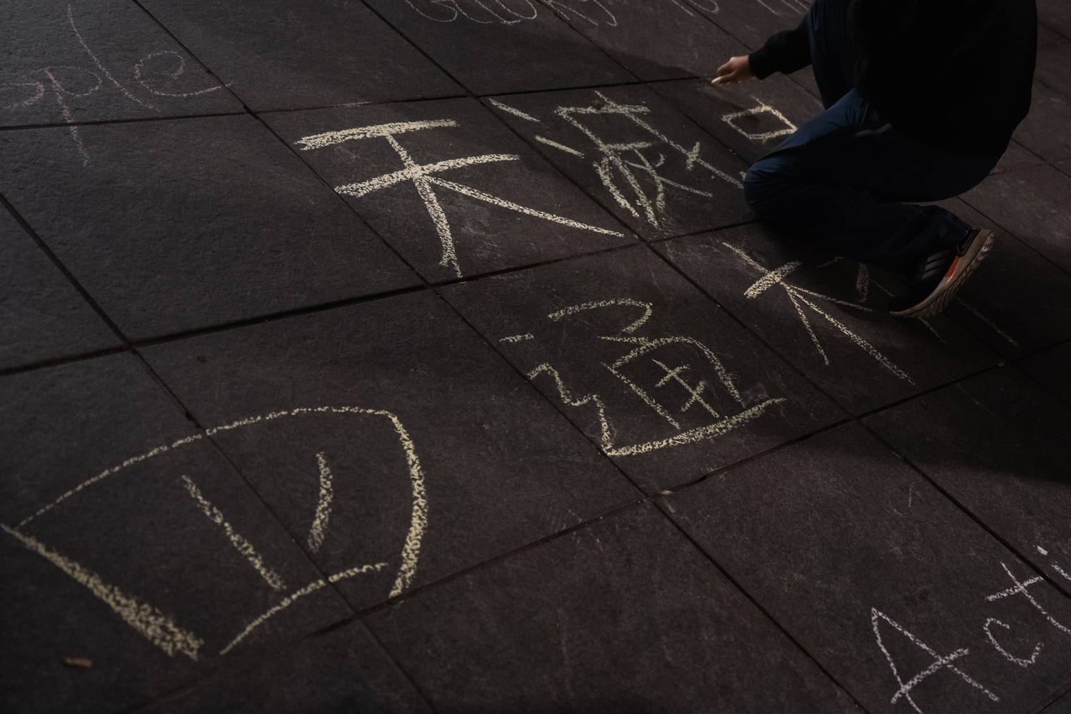 A person kneeling down on the ground while writing with yellow chalk. Next to him on the ground is chalk writing of “Sitong Bridge” in Chinese.