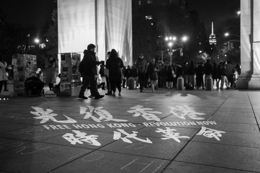 Text “FREE HONG KONG. REVOLUTION NOW” in both English and traditional Chinese projected onto the ground. In the background is a group of people standing under the Washington Square Arch and the Empire State Building. The photo is black and white.