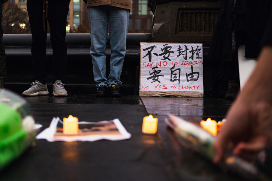A hand putting down a flower in the middle of three L.E.D. candles. In the background are a sign that reads “不要封控 要自由 Say No to Zero COVID Say Yes to Liberty” and two people standing next to the sign.