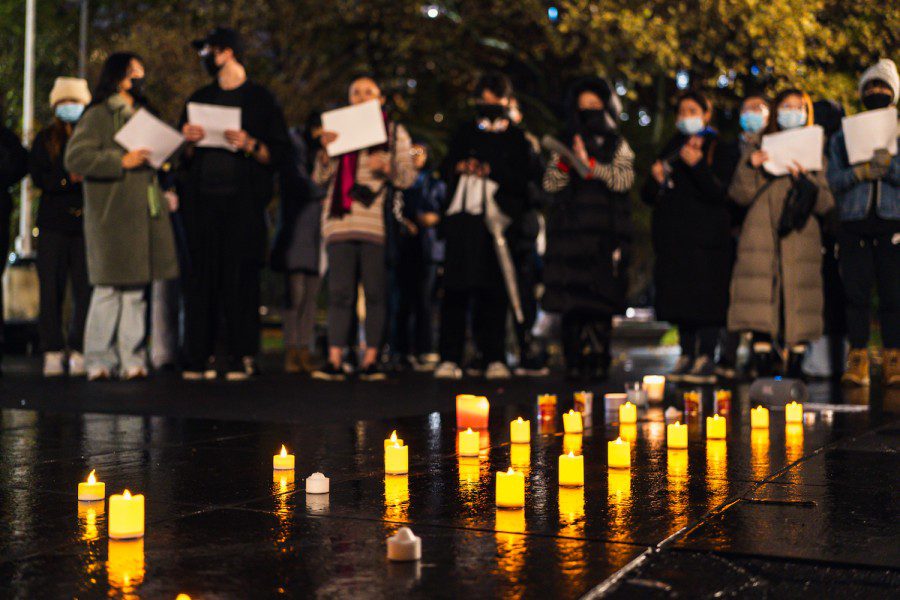 A line of candles arranged on the ground at Washington Square Park against the background of protesters standing in silence. Some protesters hold a blank sheet of white paper in their hands.