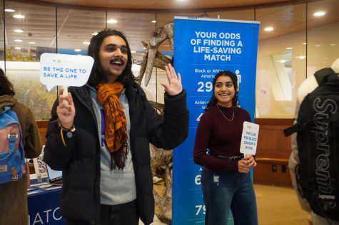 A student holds up both hands, one with a sign “BE THE ONE TO SAVE A LIFE.” Another student holds up a sign reading “THE CURE FOR BLOOD CANCER IS IN YOU.”