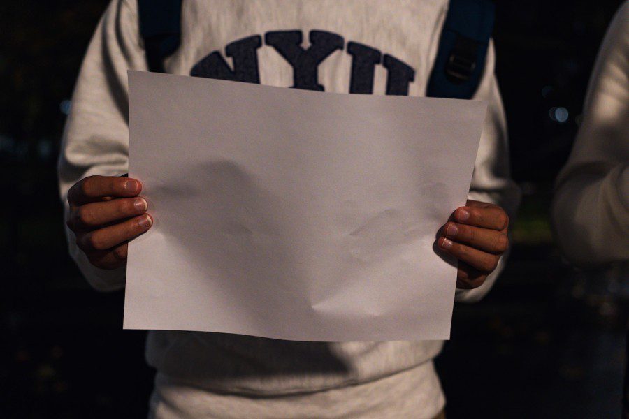 A pair of hands holding a blank sheet of white paper. The person is wearing a white sweatshirt with text “N.Y.U.” in purple.