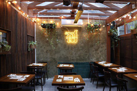 The interior of the dining area of a restaurant. In the background is a yellow neon sign. There are tables and chairs set with cutlery and menus. The walls are of a wooden texture.