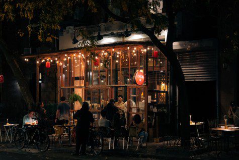 The storefront of the restaurant “Soothr” at night. The restaurant has wooden framed windows. Tables and chairs are on the sidewalk. A red logo of the restaurant sticks out from the side.