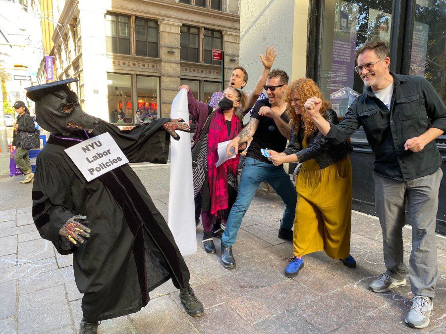 A rally attendee dressed in black as a rat with a sign that reads “N.Y.U Labor Policies” around its neck. A group of rally attendees not in costumes pretend to punch the rat.