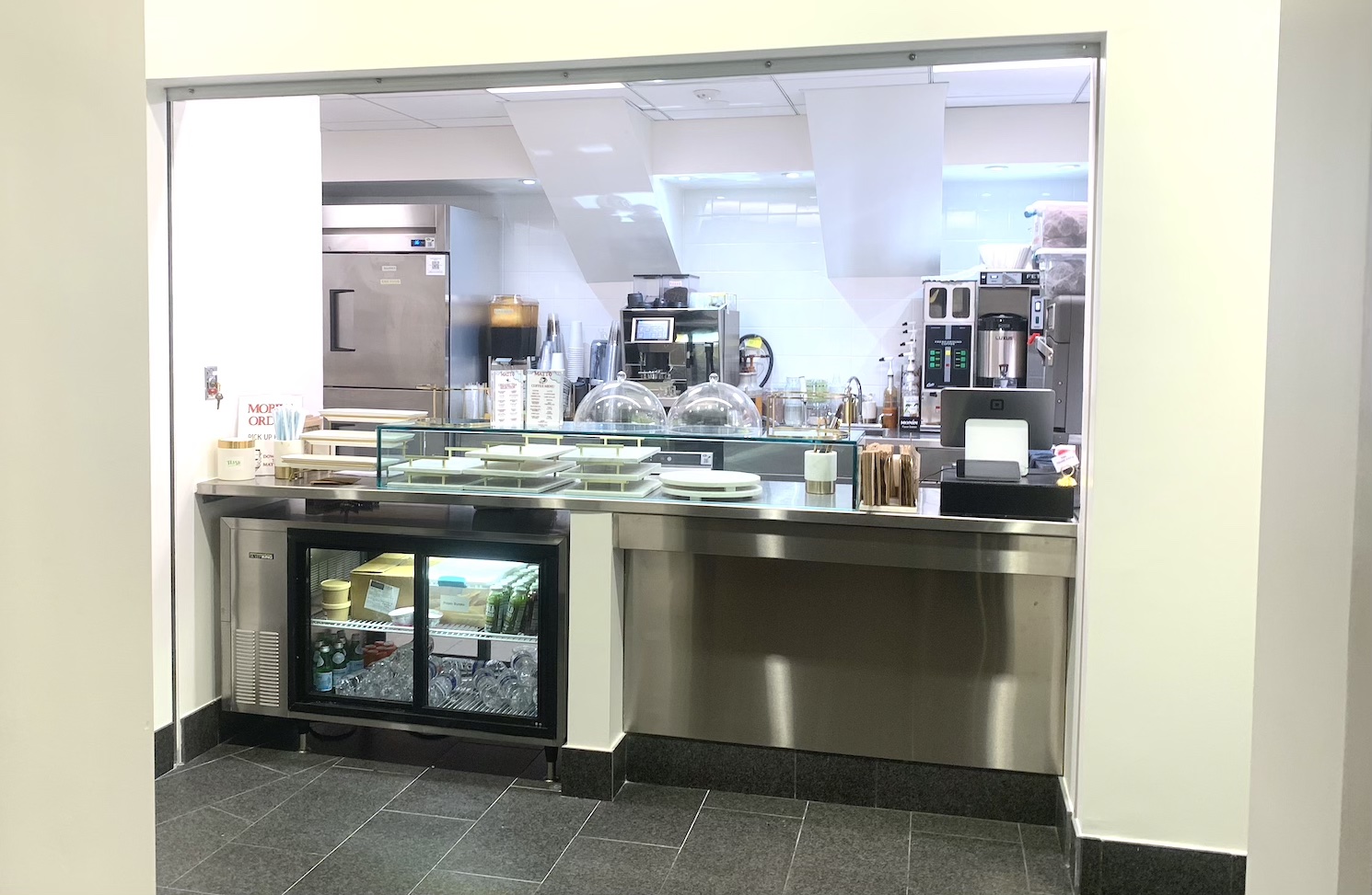 An indoor storefront with white walls, a silver metallic countertop, and a glass display case for pastries. Behind the countertop are a silver refrigerator, a coffee maker, and other equipment.
