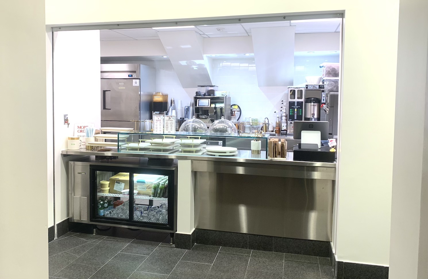 Covered showcase with white walls, silver metal top and glass baking display.  A silver refrigerator, a coffee maker and other equipment stand behind the tabletop.