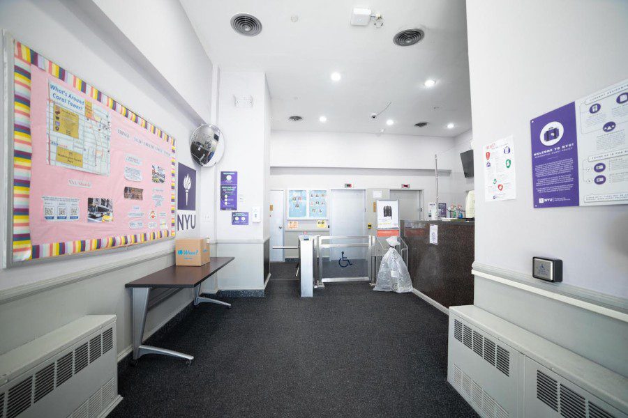 The entrance of the Coral Tower residence hall. To the left is a pink bulletin board with announcements about the dorm. In the middle is a gray gate to enter the residence hall, with two elevators in the background. To the left is a brown desk where security officers typically sit.