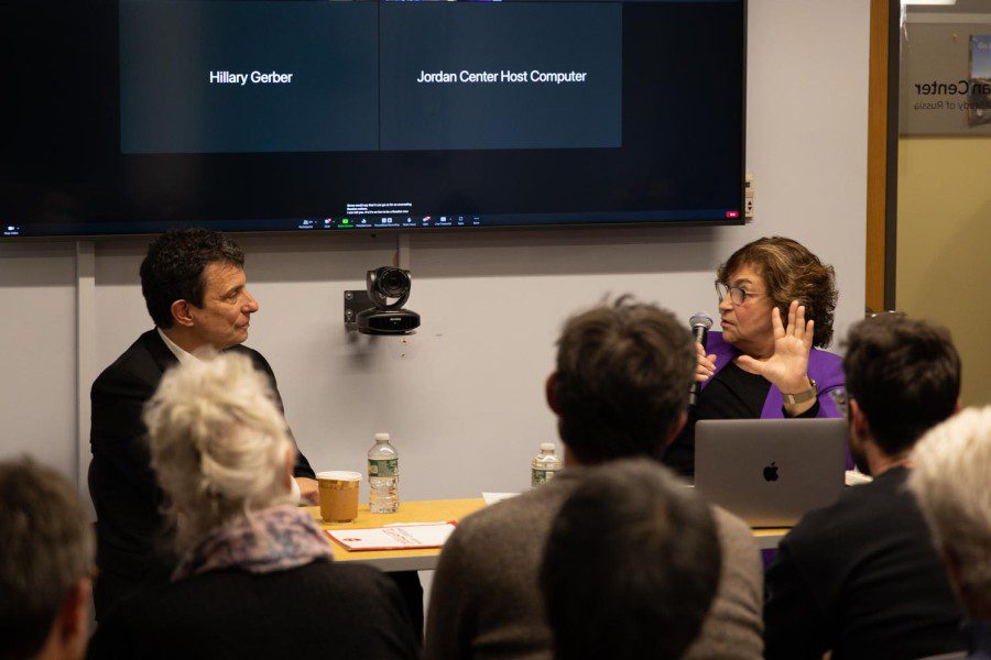 Two people sit behind a desk and a television behind them displaying a conference call. On the left is David Remnick wearing a black suit and on the right is Yevgenia Albats wearing a purple jacket. A group of people sit in front of them in a packed room.