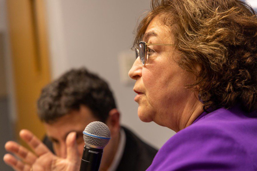 A close-up photo of Yevgenia Albats wearing a purple jacket and glasses with a gold frame. She holds a microphone while talking.