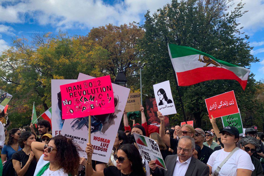 A large crowd of protestors at Washington Square Park holding up signs and Iranian flags. Some of the signs read “WOMAN LIFE FREEDOM,” “FREE IRAN,” and “IRAN 2022 REVOLUTION NO ISLAMIC! TO REPUBLIC!”