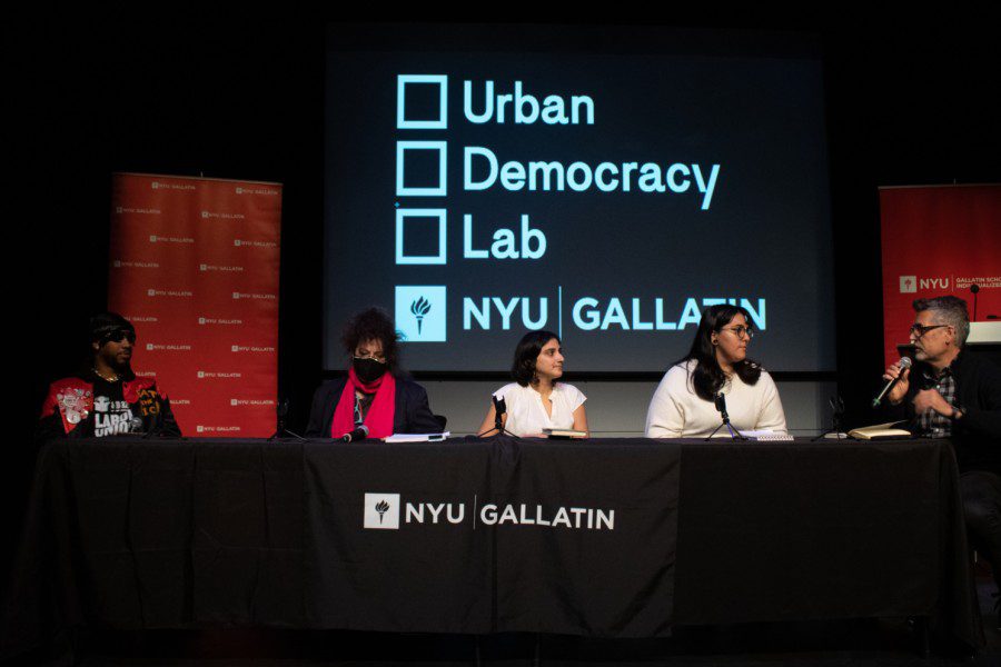 Five people sit on stage behind a black table with the text “N.Y.U. GALLATIN” written on it. Behind them is a projector screen displaying a checklist of three items: “Urban,” “Democracy” and “Lab.” The person sitting on the right holds a microphone and is speaking facing the other four people.