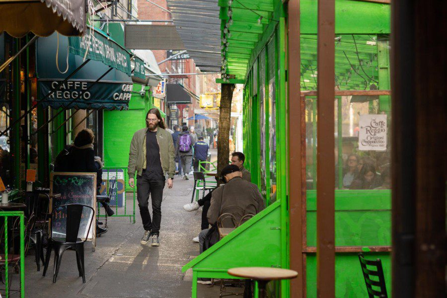 The sidewalk view of Caffe Reggio. There is an outdoor dining setup painted in green. On the other side of the sidewalk is a dark green awning with the text “CAFFE REGGIO” printed on it.