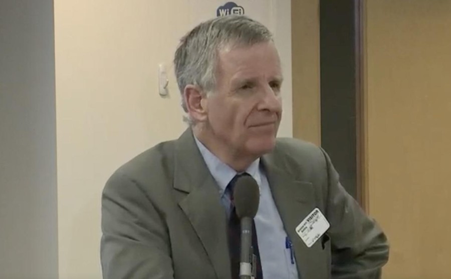 A headshot of a male with gray hair wearing a blue shirt, a dark gray tie, and a gray blazer as he talks into a microphone.
