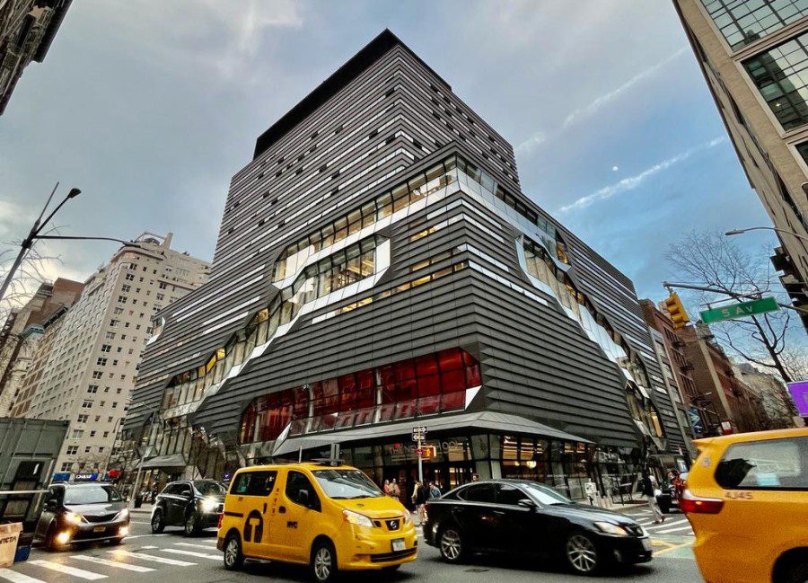 A gray, modern building on the corner of a busy intersection on Fifth Avenue. Yellow taxi cabs and other cars pass by.