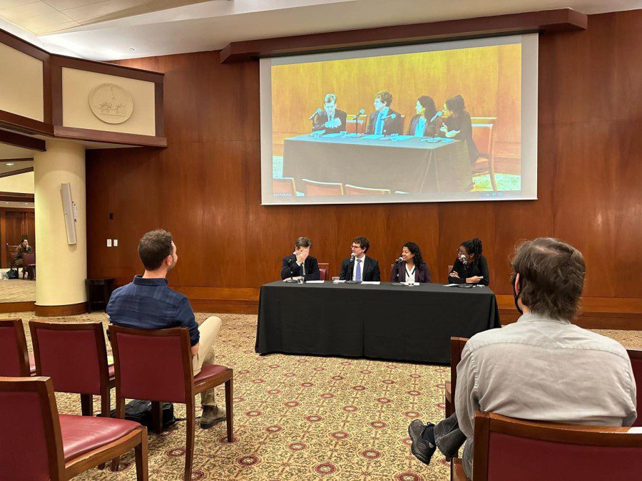 Brad Hoylman, Noah Rosenblum, Alice Fontier, and Tasleemah Lawal sit at a panel discussion event. All four are wearing formal attire, including dark-colored blazers. Above the panelists is a screen projecting a video recording of the panel. In front of the panelists are members of the audience sitting in chairs listening to the discussion.