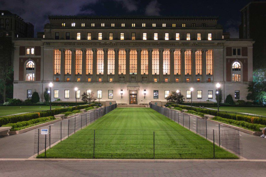 An exterior view of Columbia University’s Butler Library at night. The library has marble ionic columns and engravings of the names of various Greek thinkers on the beams.