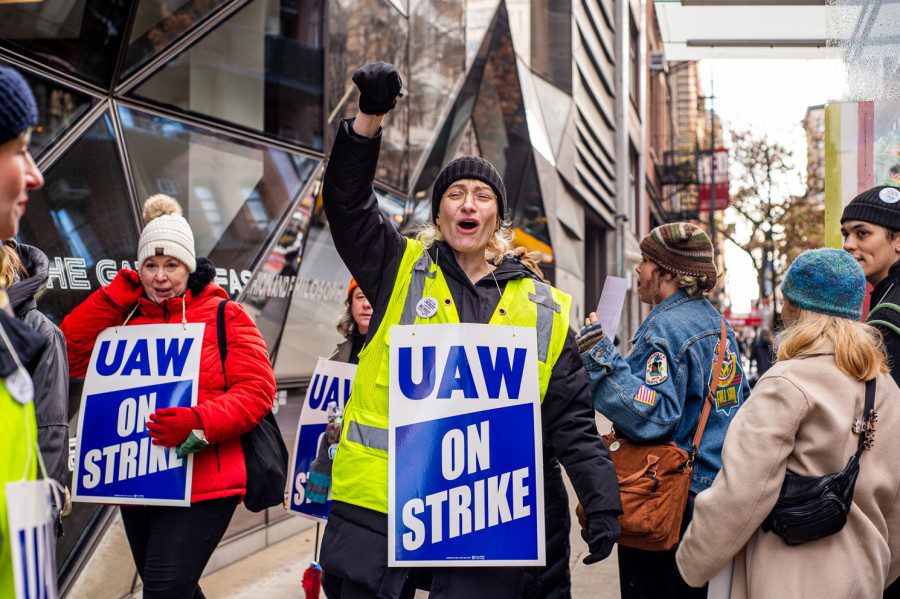 A part-time faculty member on strike. They hold up their fist and carry a sign that reads U.A.W. ON STRIKE. Behind them are other striking faculty members.