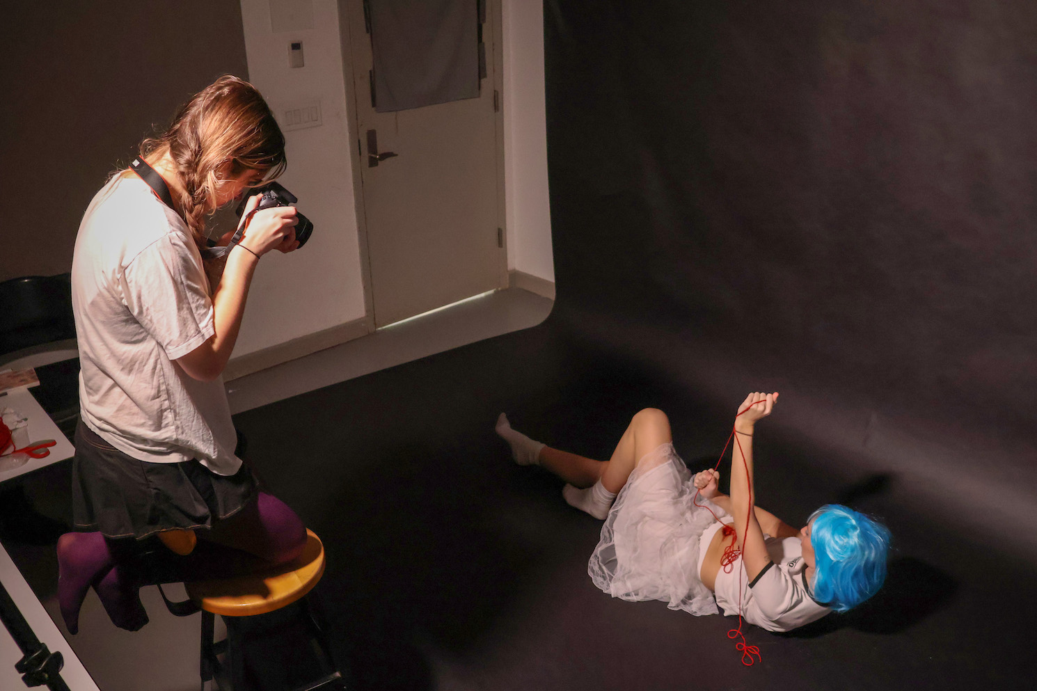 Photograph of a female college student with braided blond hair wearing a white t-shirt, a black skirt, maroon leggings, and gold bracelets holding a DSLR camera taking a photo of a girl laying on the ground in front of a black backdrop wearing a short blue wig, white crop top, white fluffy skirt, holding red string in her hands.