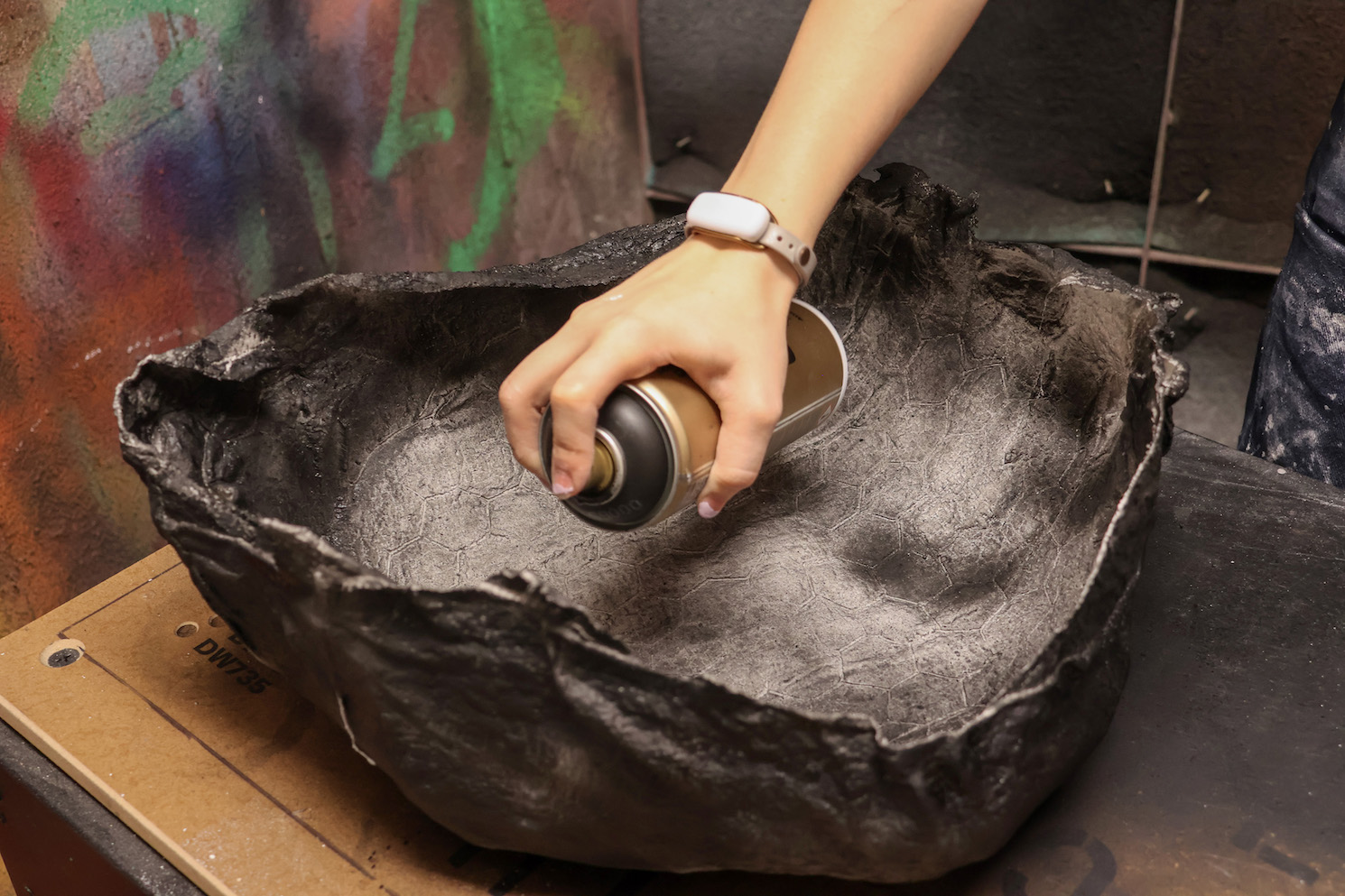 A hand wearing a white watch holds a bottle of black spray paint and sprays the paint onto a black bowl.