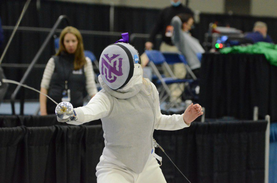 A fencing athlete wearing a white fencing suit and a helmet with the N.Y.U. logo printed on it extends their fencing épée forward.