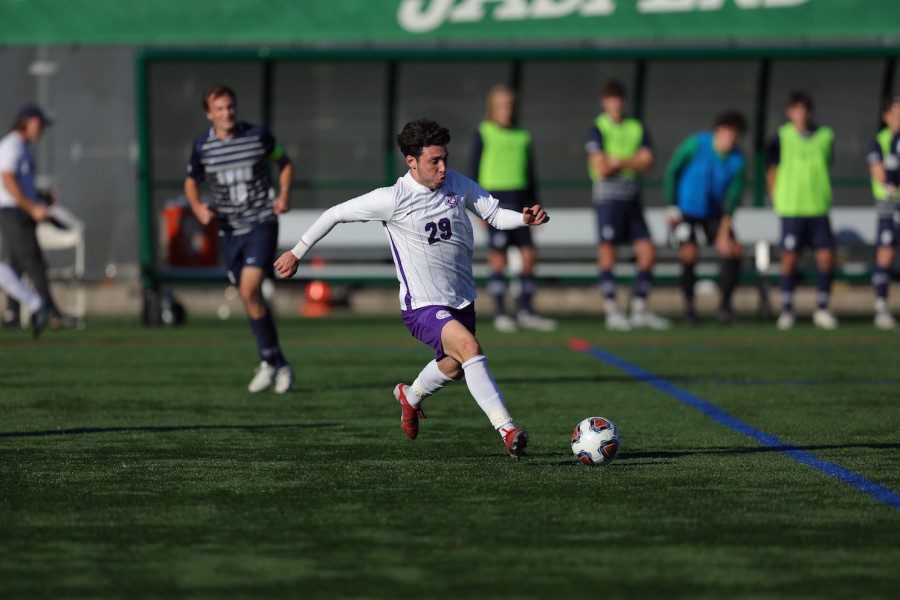 Joe Leslie, player number 29, kicks a ball down the field wearing a white N.Y.U. jersey, purple shorts, and red soccer shoes.