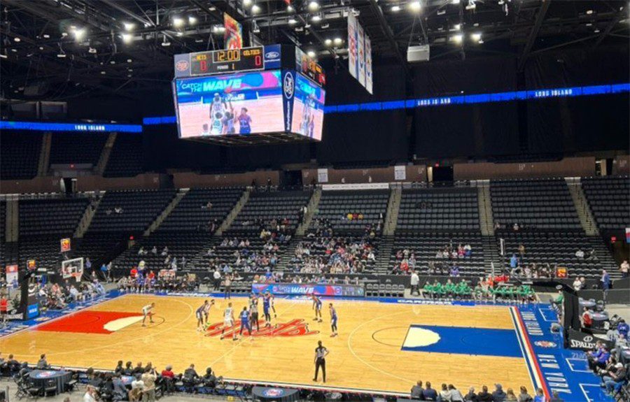 The interior of the Nassau Veterans Memorial Coliseum. In the center is a basketball court with players practicing. Around the court are numerous rows of black seats with fans sitting and standing.