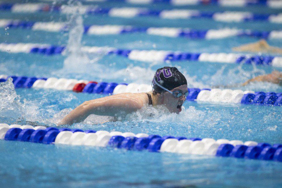 NYU swimmer Caitlin Marshall swims in butterfly style in a pool. Marshall wears a dark blue swimsuit, a dark swim cap with purple text outlined in white reading N.Y.U,and a pair of green reflective goggles.